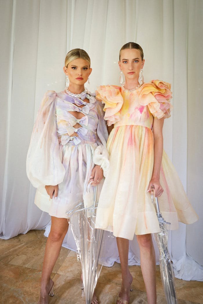 Two Caucasian women in elegant dresses pose indoors with translucent umbrellas.